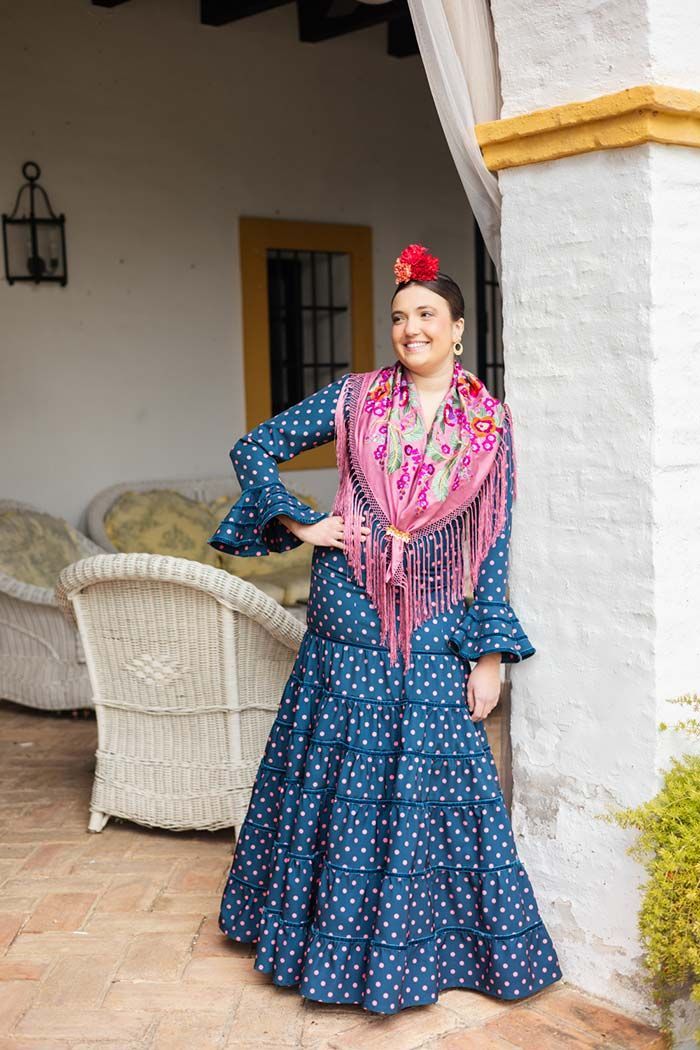 Mujer con vestido azul de lunares y chal rosa posando en un patio junto a muebles de mimbre blancos.
