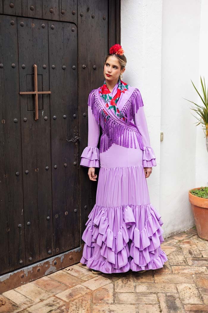 Una persona vestida con un traje de estilo flamenco color lavanda está de pie junto a una puerta de madera oscura y una pared blanca.