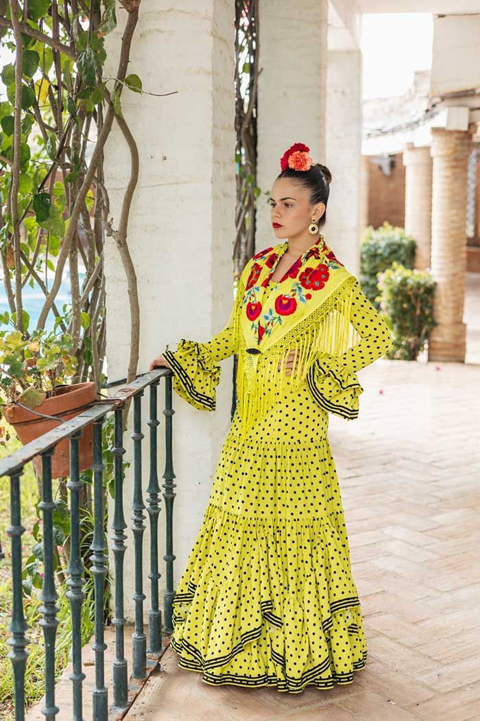 Una persona con un vestido amarillo brillante con volantes y flores rojas está de pie junto a una barandilla en un patio soleado.