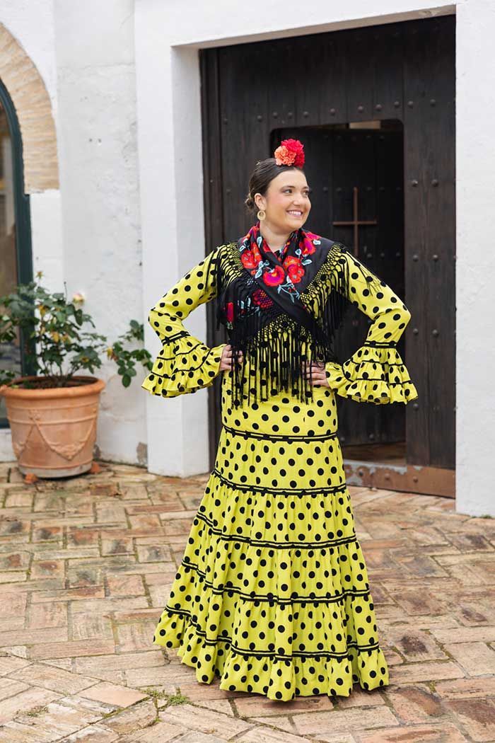 Una persona con un vestido flamenco amarillo de lunares posa en un patio, con las manos en las caderas, junto a una puerta.