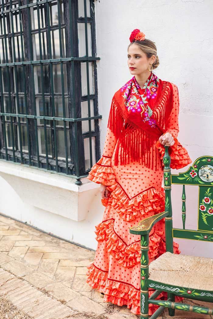Mujer con traje tradicional rojo de pie junto a una ventana y un banco verde al aire libre.