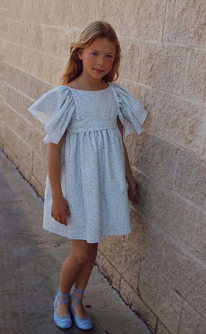 Niña con un vestido azul claro de pie junto a un muro de piedra al aire libre.