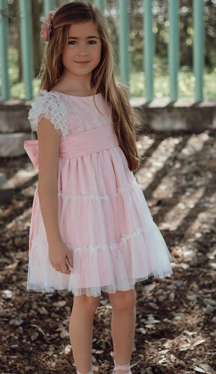 Niña pequeña con un vestido rosa de pie al aire libre cerca de una valla, con la luz del sol y las hojas caídas a su alrededor.