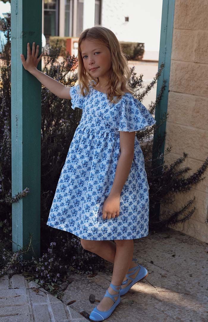 Una chica con un vestido azul estampado de flores está de pie junto a una puerta, con la mano apoyada en la pared y sandalias azules.