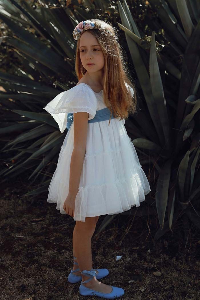 Niña con vestido blanco y zapatos azules de pie frente a grandes hojas tropicales.