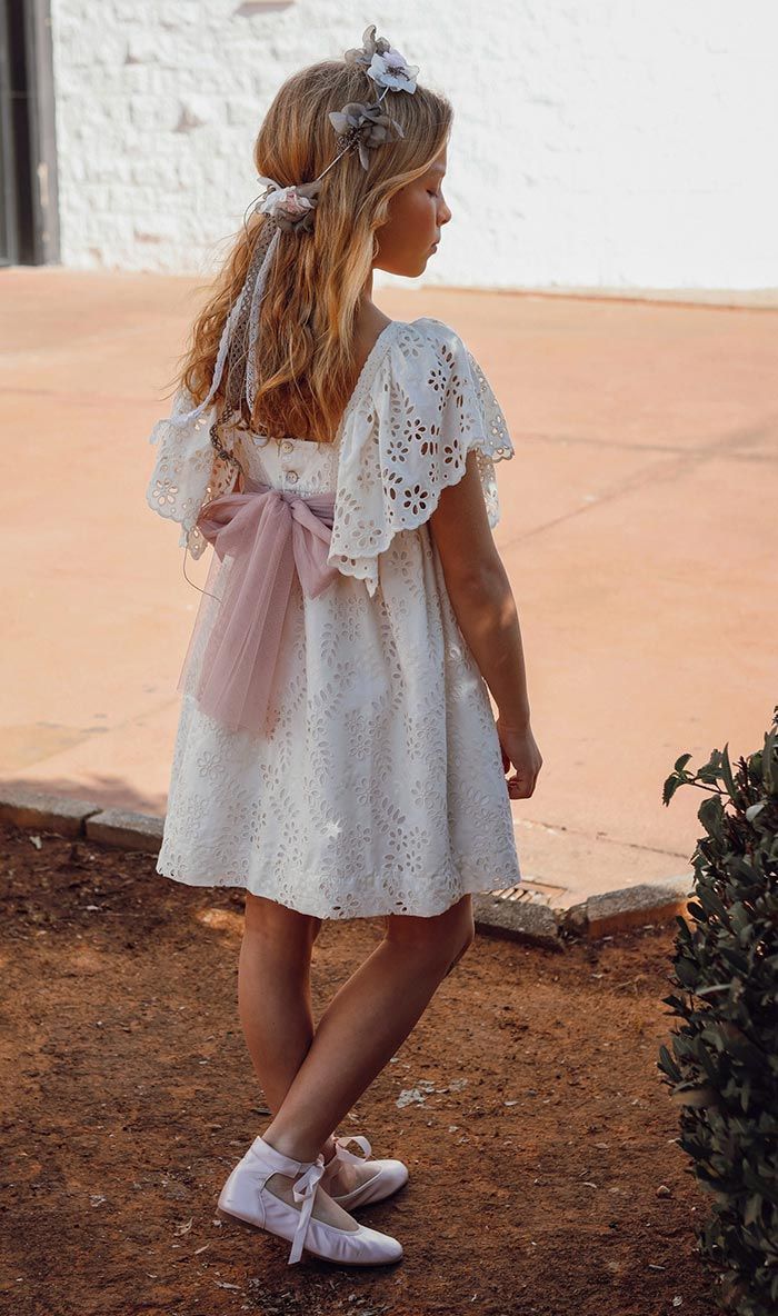 Niña con vestido blanco y sandalias de pie al aire libre en un patio de ladrillos, vista desde atrás.