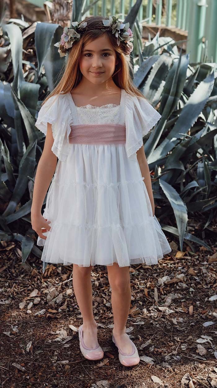 Niña con vestido blanco y zapatos rosas de pie al aire libre frente a plantas de agave.