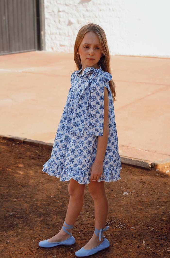 Una niña con un vestido azul estampado de flores y zapatos está de pie al aire libre en una zona pavimentada cerca de un muro.