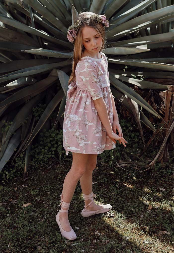 Niña con vestido rosa y corona de flores de pie al aire libre frente a grandes plantas verdes puntiagudas.