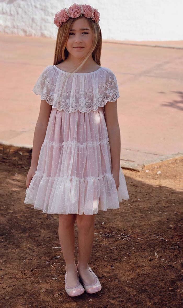 Niña con un vestido rosa de encaje y una corona de flores de pie al aire libre sobre tierra cerca de una zona pavimentada.