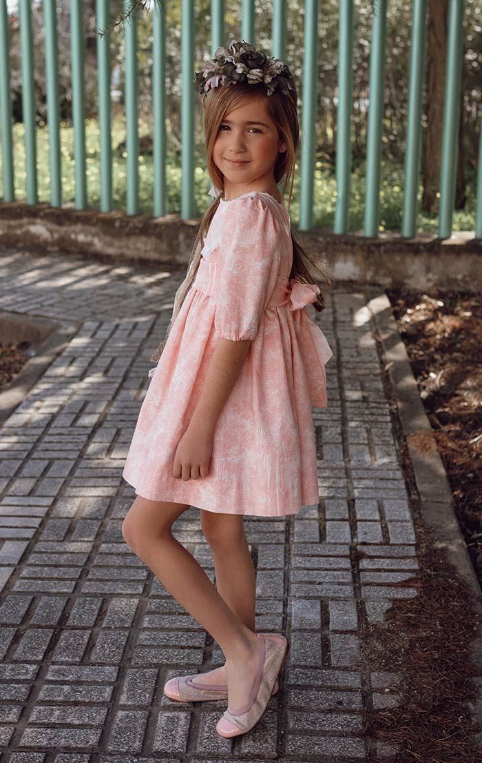 Niña con un vestido rosa posando en un camino de ladrillos junto a una valla metálica.