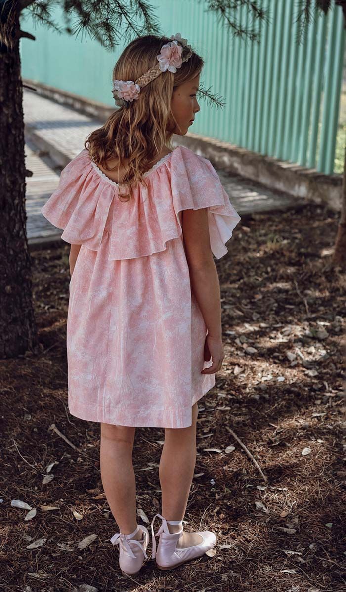 Niña con vestido rosa y diadema floral de pie al aire libre junto a una pared de metal corrugado.
