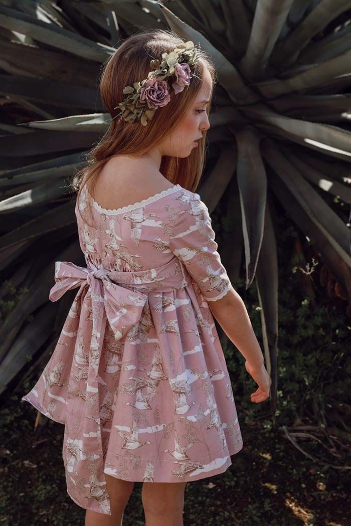Niña pequeña con vestido de encaje rosa y corona de flores de pie entre grandes plantas de color verde oscuro.