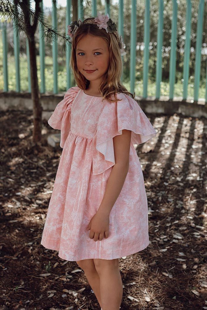 Una niña con un vestido rosa con volantes está de pie al aire libre, cerca de una valla, bajo la luz del sol moteada.