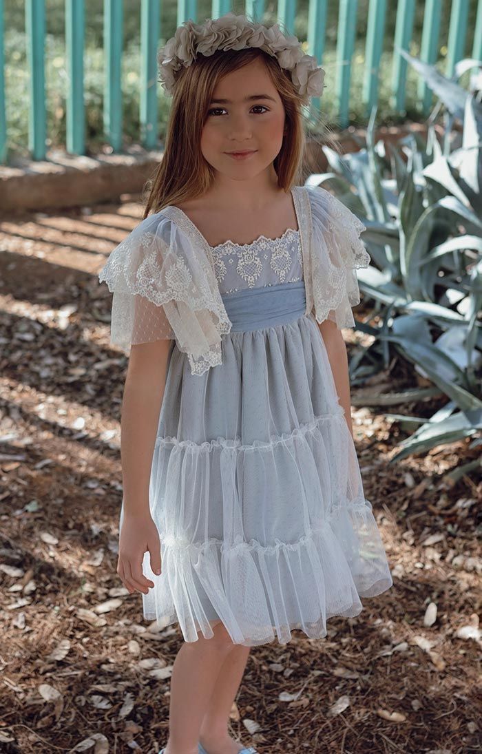 Niña con un vestido blanco de encaje de pie al aire libre sobre virutas de madera cerca de una valla verde y plantas.