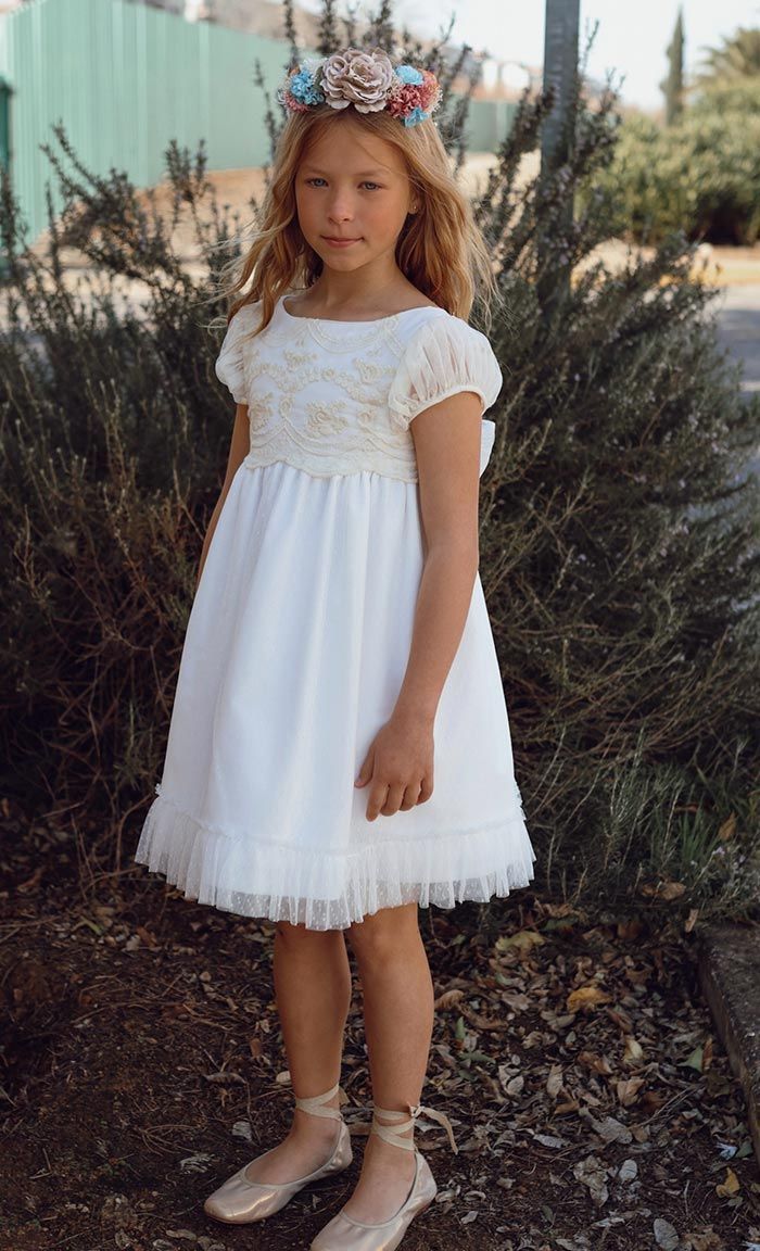 Niña con vestido blanco y corona de flores de pie al aire libre frente a unos arbustos altos.