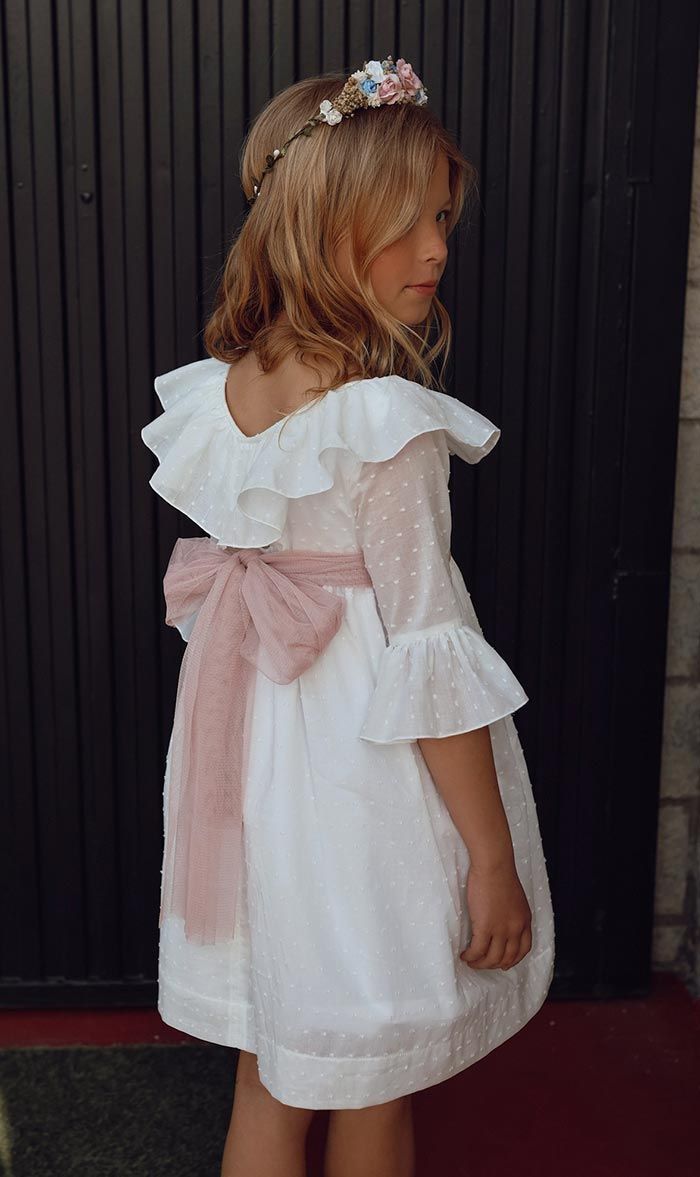 Niña con un vestido blanco, un lazo rosa y una diadema floral, de perfil contra una cortina oscura.