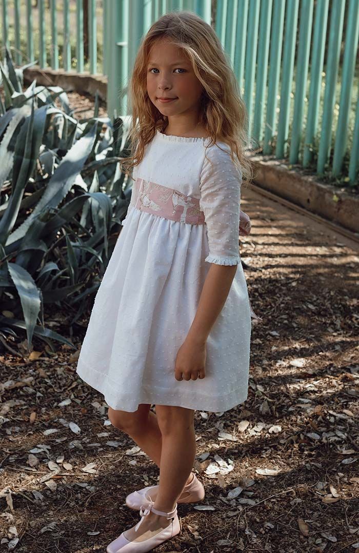 Una joven con un vestido blanco está de pie al aire libre junto a una valla verde, con árboles y hojas caídas cerca.