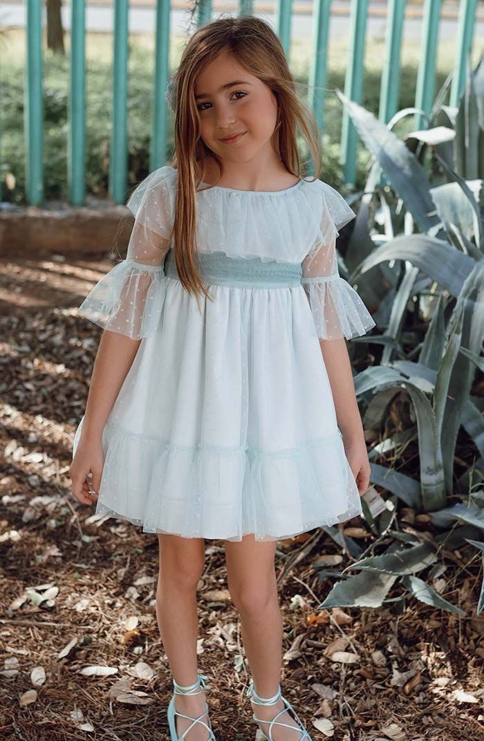 Niña pequeña con un vestido blanco de pie al aire libre junto a una valla y plantas.