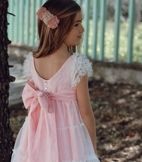 Niña con un vestido rosa con un lazo, de pie al aire libre cerca de una valla.
