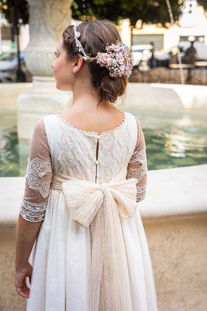 Mujer con un vestido blanco de encaje y un tocado floral, de pie junto a una fuente de espaldas.