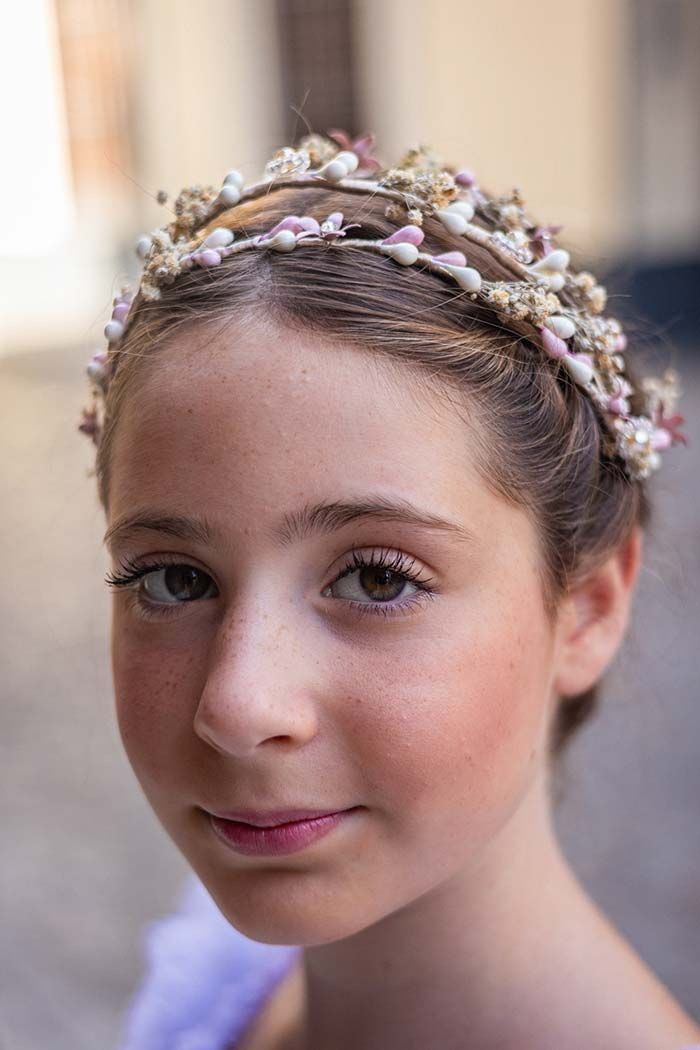 Retrato en primer plano de una niña con una diadema floral, mirando a la cámara con una dulce sonrisa.