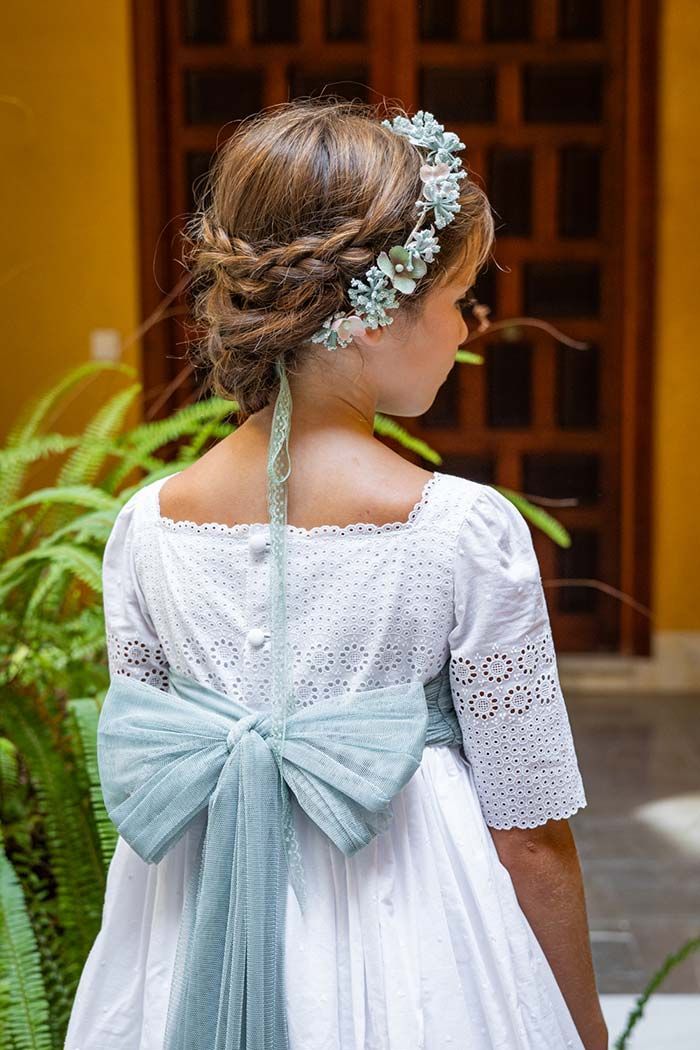 Niña con un vestido blanco y un gran lazo azul claro, vista de espaldas en un patio ajardinado.