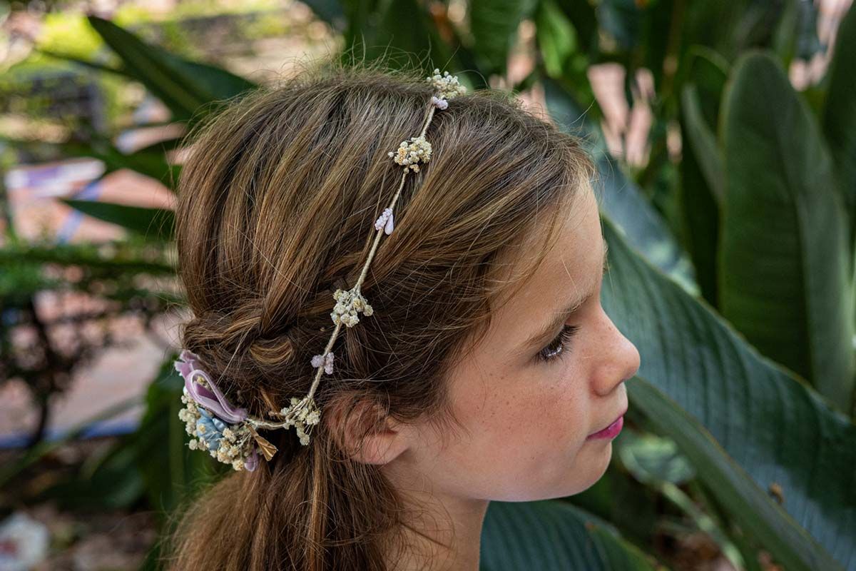 Niña de perfil con corona de flores en el cabello, de pie entre grandes hojas verdes.