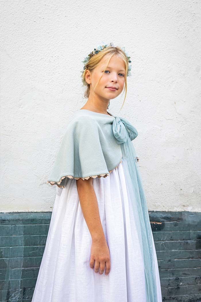 Una niña con un vestido azul claro y una diadema floral está de pie frente a una pared blanca y una base de ladrillo oscuro.