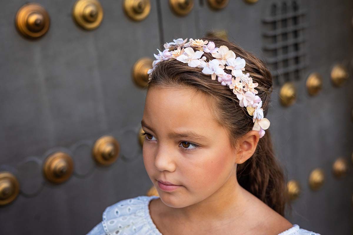 Niña con diadema floral, mirando de reojo contra una pared gris con pomos dorados.