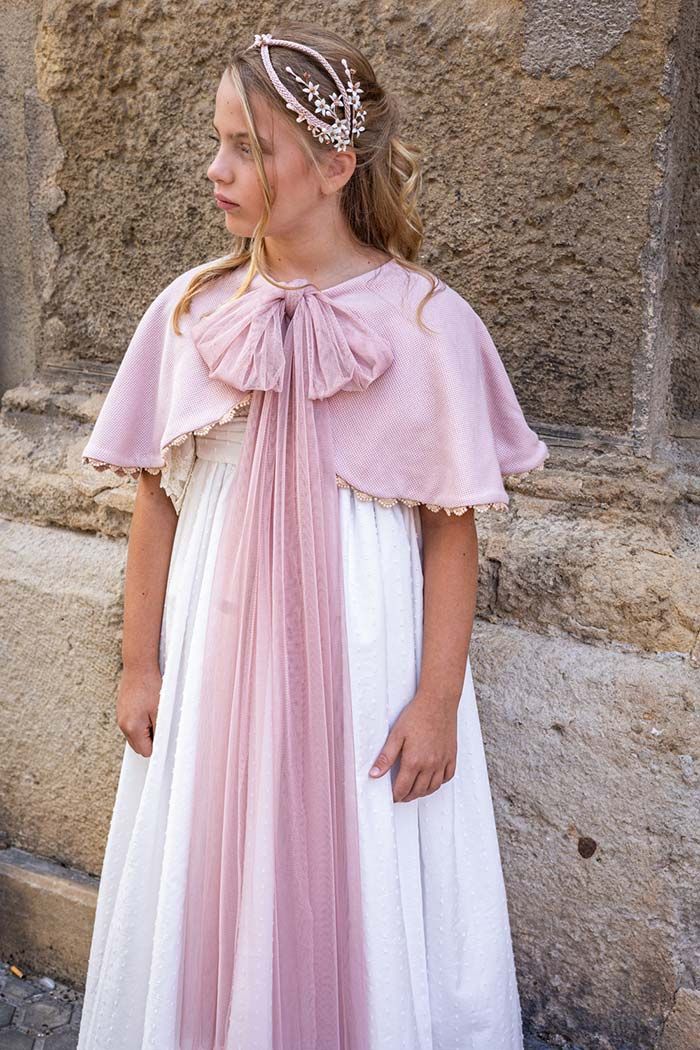 Niña pequeña con un vestido blanco y una capa rosa, de pie junto a un muro de piedra.