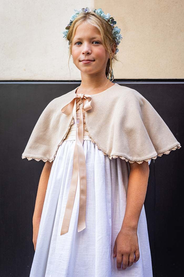 Una niña vestida con un vestido blanco y una capa beige está de pie junto a una pared lisa.