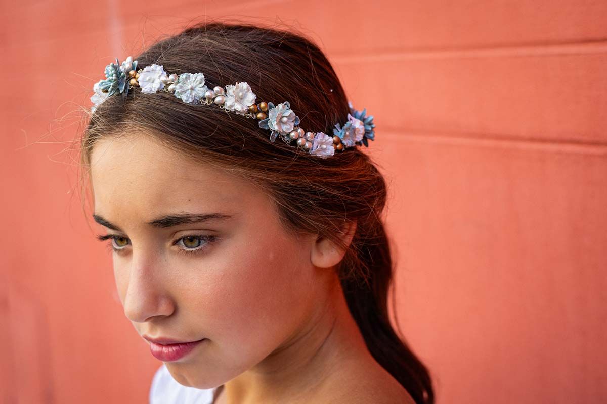 Retrato de una niña con una diadema floral contra una pared de color coral, mirando hacia abajo pensativa.