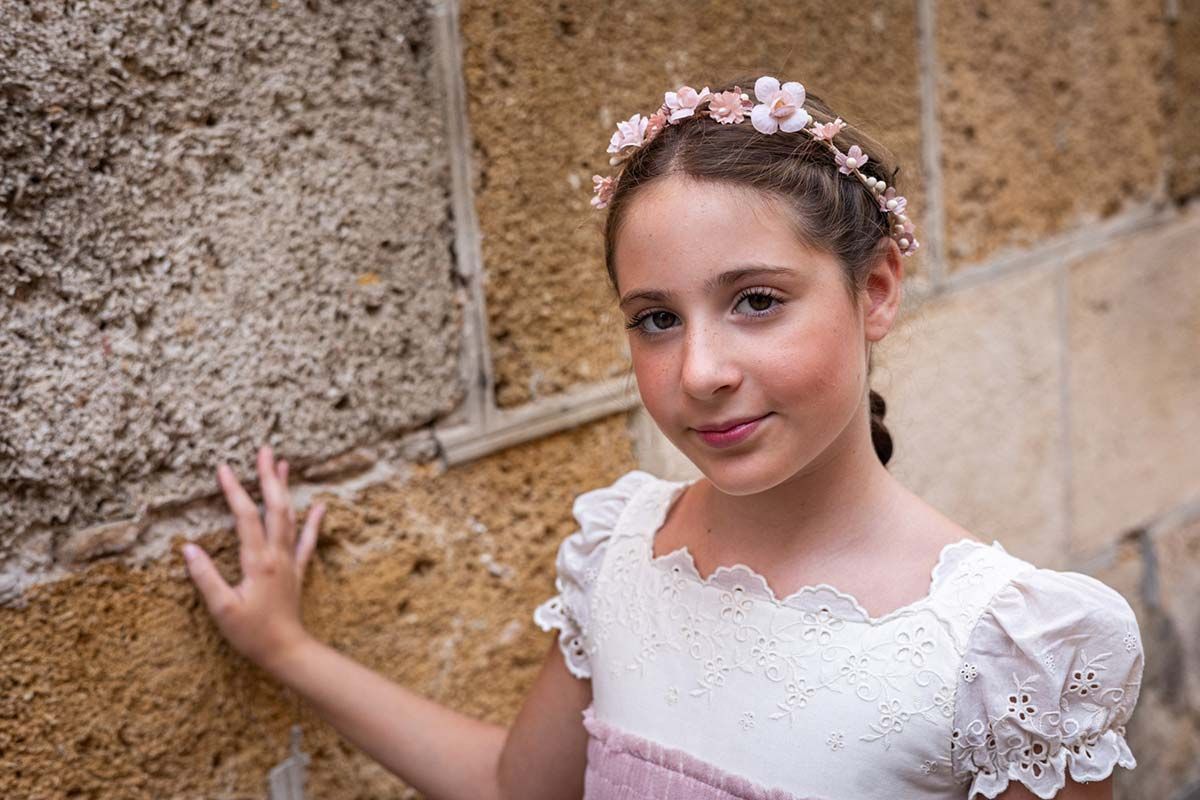 Mujer joven con vestido blanco y corona de flores, apoyada contra un muro de piedra y sonriendo dulcemente.