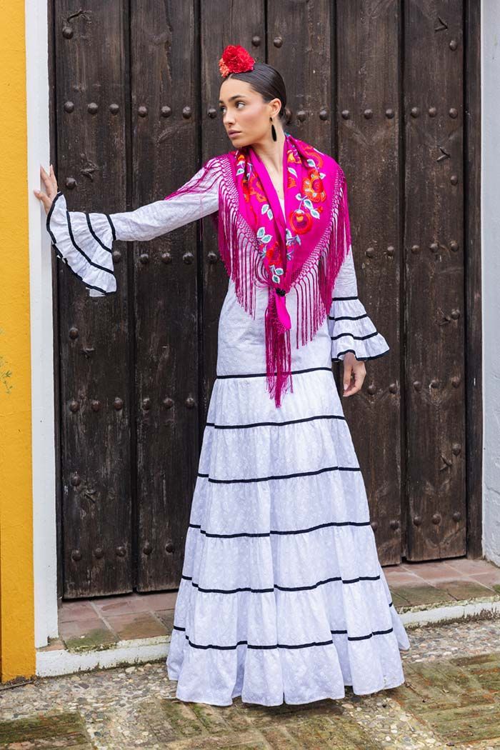 Mujer con vestido blanco de rayas negras y chal rosa de pie junto a una puerta de madera.