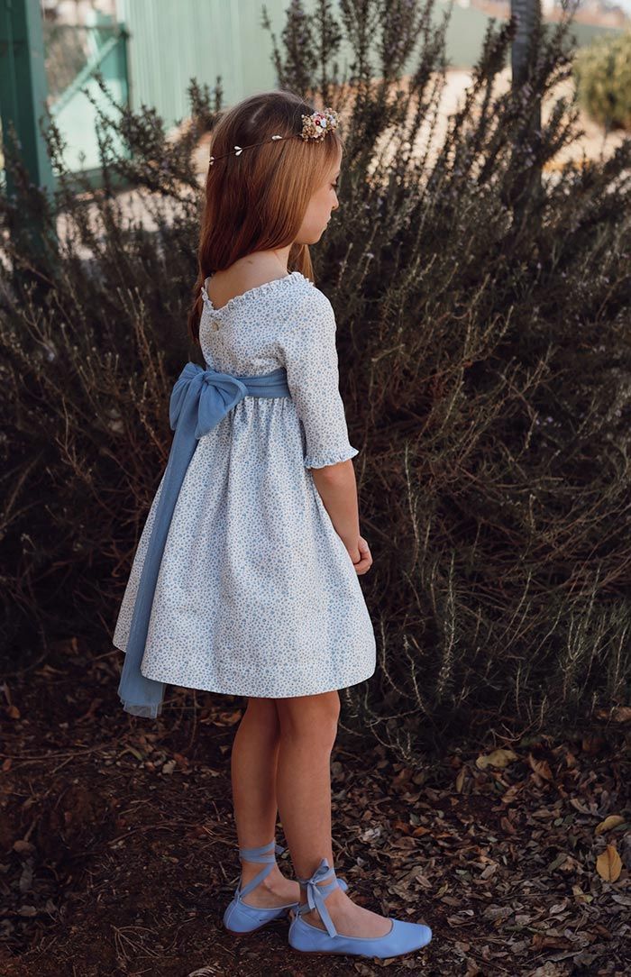 Niña con vestido y zapatos azul claro de pie junto a unos arbustos altos al aire libre.