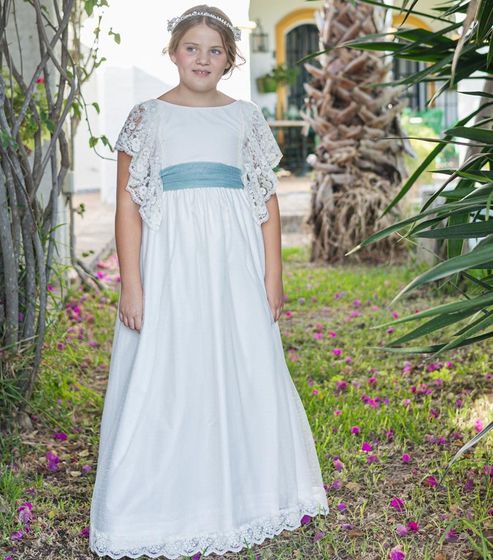 Mujer con vestido blanco y fajín azul de pie en un sendero de jardín, rodeada de palmeras y flores.