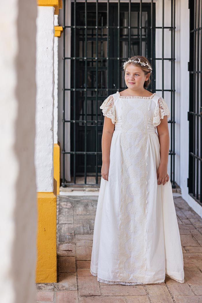 Mujer con un vestido de encaje blanco de pie en una puerta con rejas de cristal y paredes ribeteadas de amarillo.