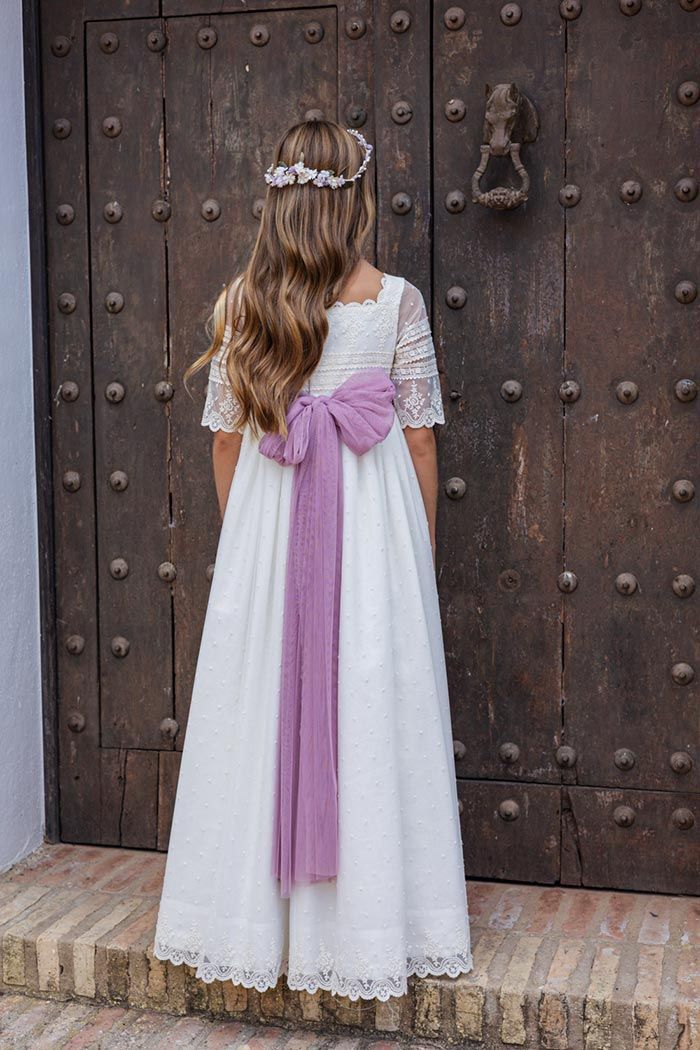 Niña con un vestido blanco y una faja lila de pie frente a una gran puerta de madera.