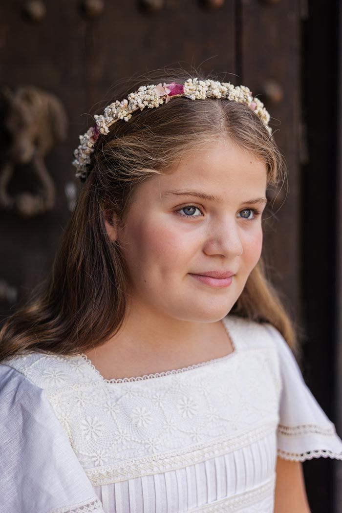 Niña con un vestido blanco y una diadema floral, sonriendo dulcemente en el interior de una casa junto a una puerta oscura.