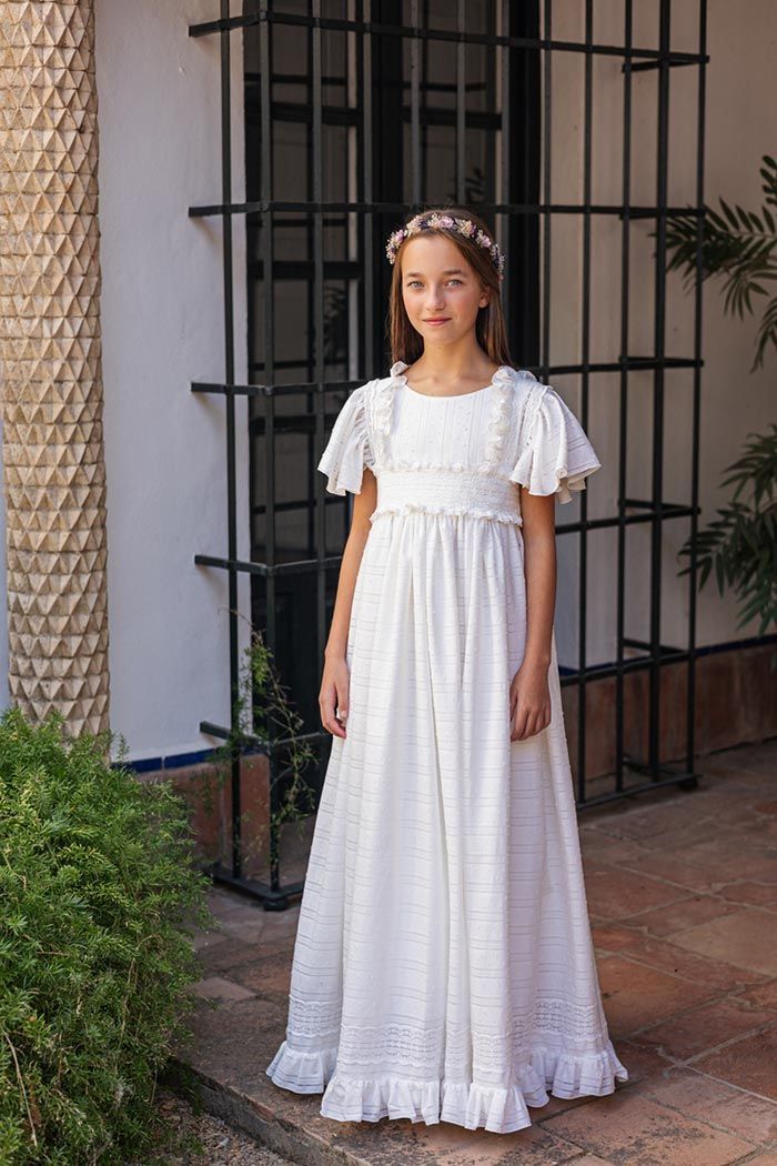 Niña con un vestido blanco largo de pie al aire libre junto a un muro y vegetación.