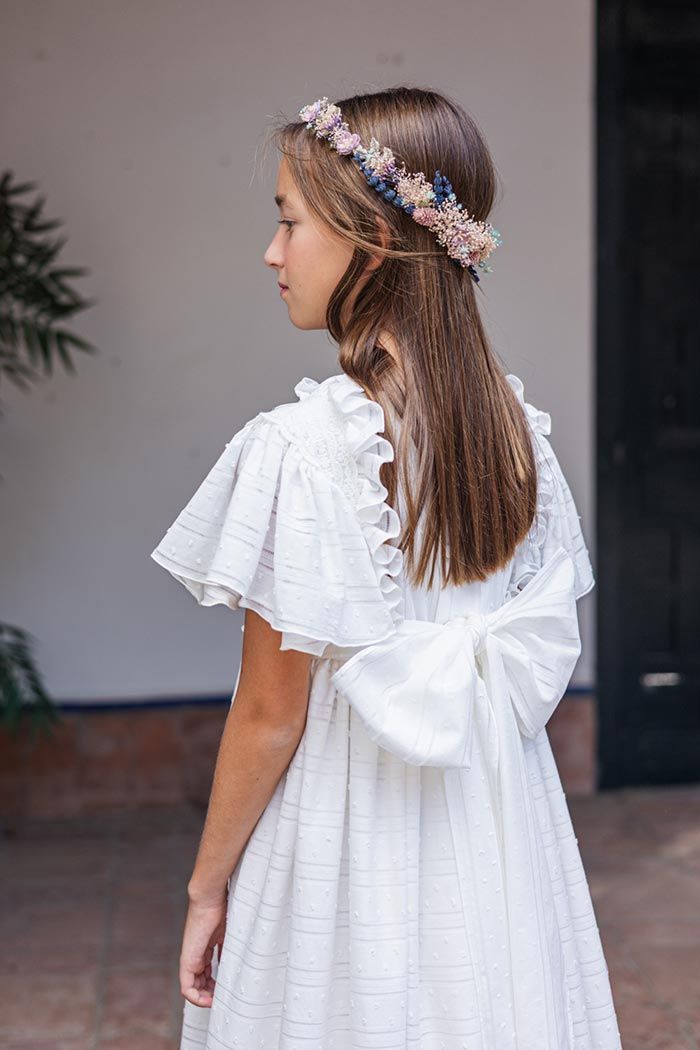 Niña con vestido blanco y diadema floral, vista de espaldas en una puerta.