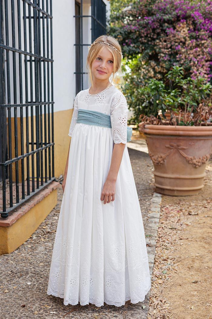 Niña con vestido blanco de pie al aire libre junto a un muro y macetas con flores.