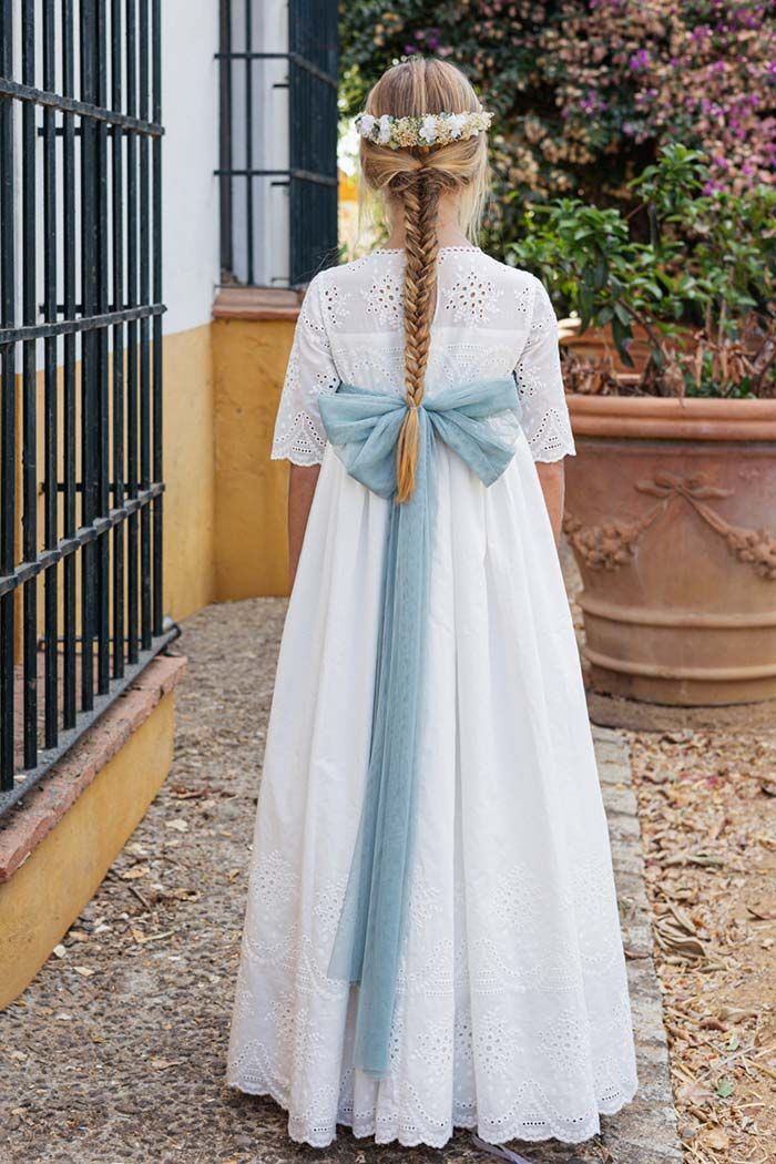 Niña con vestido blanco de encaje y lazo azul de pie al aire libre junto a un edificio y macetas.