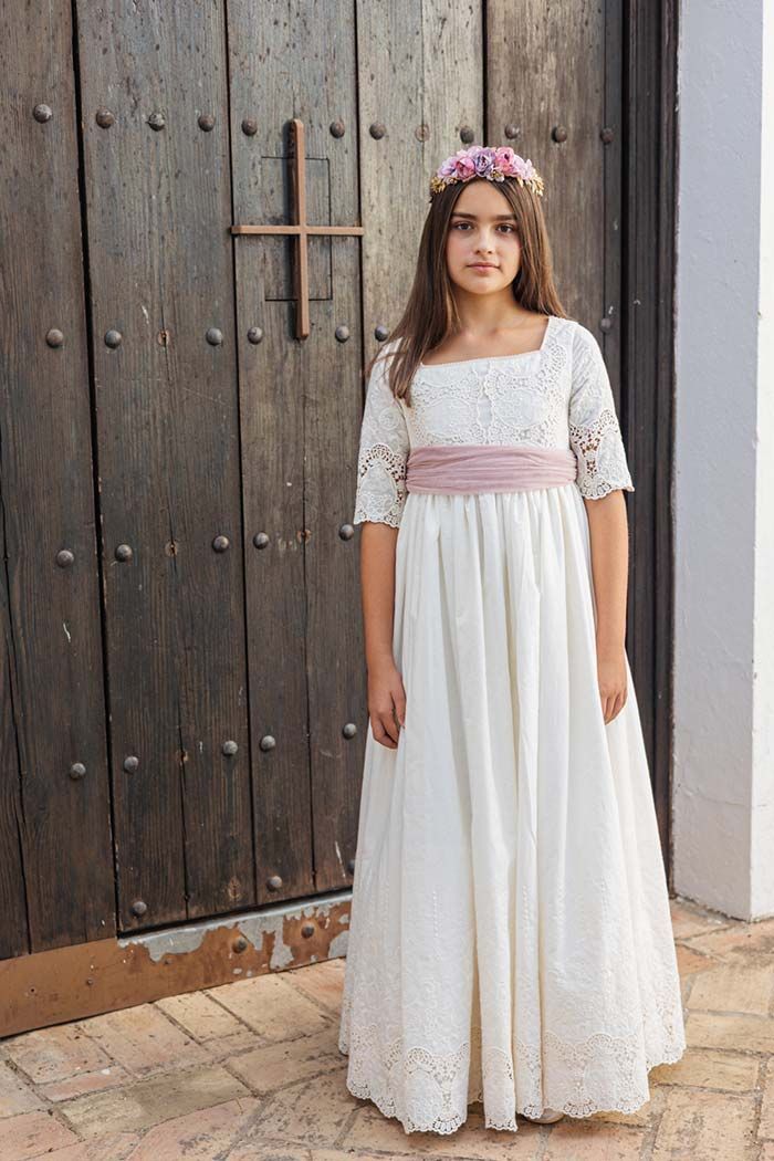 Niña con un vestido blanco de encaje, una faja rosa y una corona de flores, de pie junto a una puerta de madera.