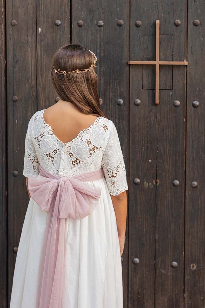 Una mujer con un vestido blanco de encaje y un lazo rosa está de pie frente a una puerta de madera con una manija en forma de cruz.