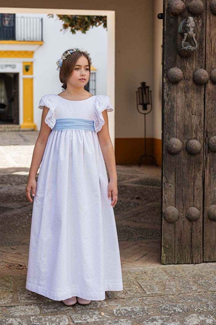 Mujer con un vestido blanco y azul claro de pie junto a una puerta rústica de madera al aire libre.