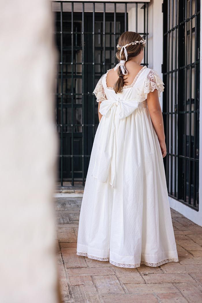 Mujer con un vestido blanco de pie junto a una puerta con verja, vista desde atrás.