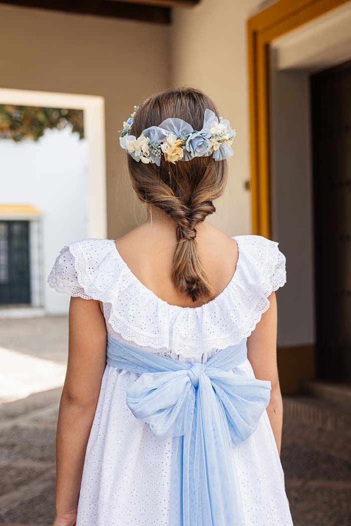 Niña con un vestido azul claro y una corona de flores en el pelo, vista de espaldas en una puerta.