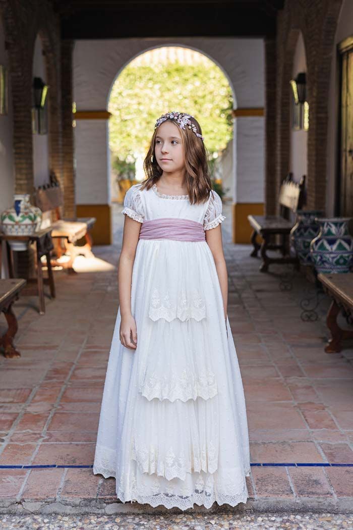 Una joven con un vestido blanco está de pie en un pasillo de ladrillos con una abertura arqueada iluminada por el sol detrás de ella.